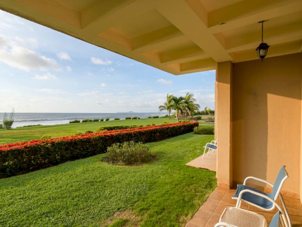 A patio overlooking a lush green lawn, colorful hedges, palm trees, and a distant ocean view, with chairs set for relaxation.