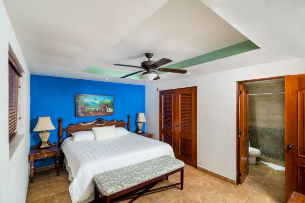 A cozy bedroom with a large bed, blue accent wall, ceiling fan, bedside tables, lamps, art, wooden doors, a bench, and a bathroom in the corner ending the sentence.