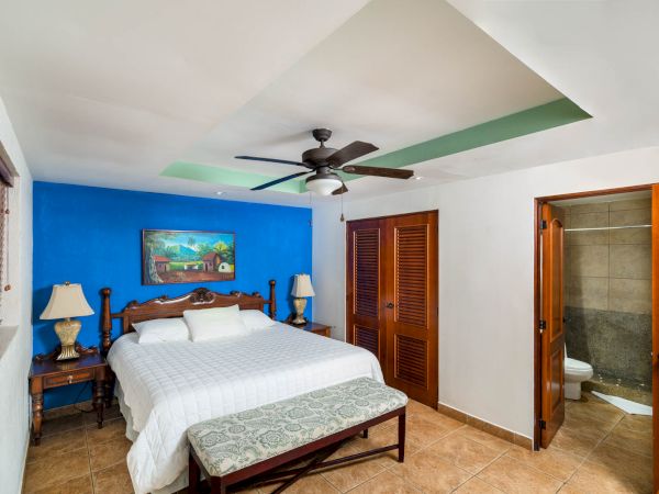 A cozy bedroom with a large bed, blue accent wall, ceiling fan, bedside tables, lamps, art, wooden doors, a bench, and a bathroom in the corner ending the sentence.