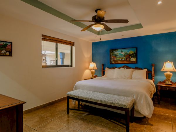 A cozy bedroom features a large bed, blue accent wall, ceiling fan, wooden furniture, bedside lamps, bench, and paintings, under warm lighting.