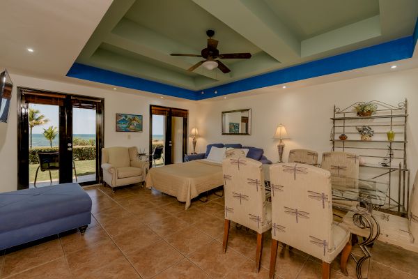 The image shows a spacious room with a king-sized bed, sitting area, dining table, ceiling fan, and large windows offering a view, with decor and furnishings.