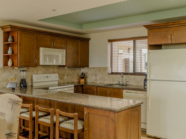 A kitchen features wooden cabinetry, a marble countertop, white appliances including a refrigerator and stove, a sink, and three bar stools.