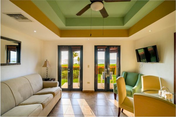 The image shows a bright, well-lit room with a sofa, chairs, a table, a TV, a ceiling fan, and a view of a garden through glass doors.