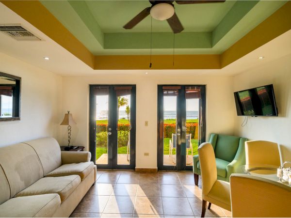The image shows a bright, well-lit room with a sofa, chairs, a table, a TV, a ceiling fan, and a view of a garden through glass doors.