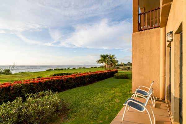 A scenic ocean view from a patio with two chairs, overlooking a well-manicured lawn and garden with palm trees under a partly cloudy sky.