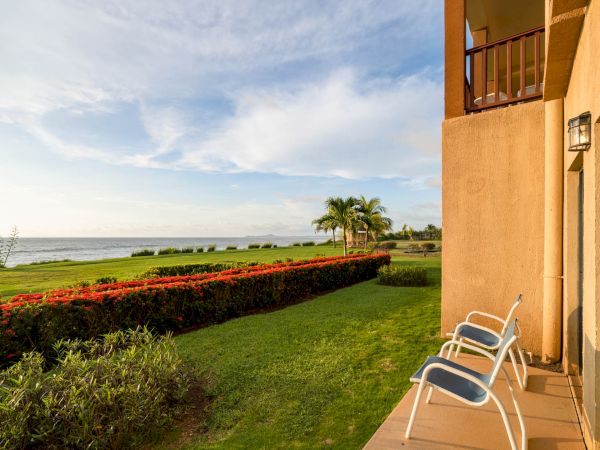 A scenic ocean view from a patio with two chairs, overlooking a well-manicured lawn and garden with palm trees under a partly cloudy sky.