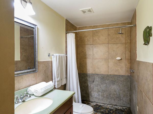 This image shows a bathroom with a sink, mirror, toilet, and a shower area with tiles. Towels are hanging near the sink, and there's a bath mat.