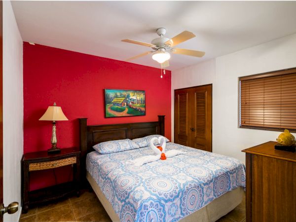 This image shows a cozy bedroom with a red accent wall, double bed, ceiling fan, side table with lamp, dresser, and painting above the bed.