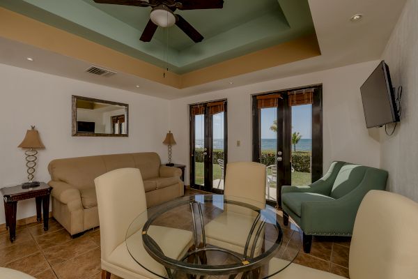 A living room with a sofa, chairs, a glass table, lamps, a ceiling fan, a wall-mounted TV, and doors leading outside with a view.