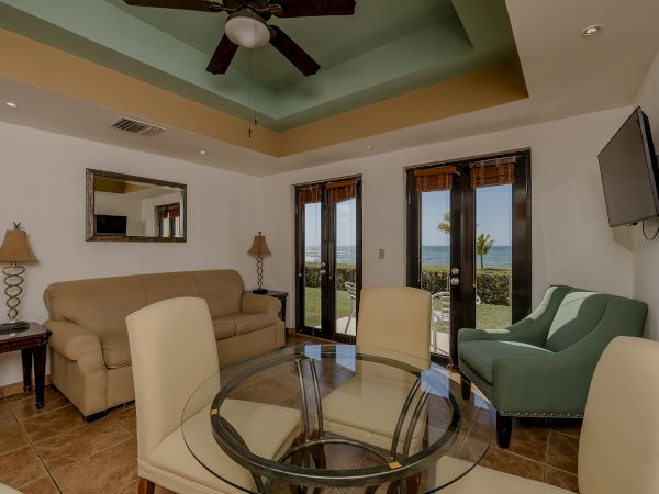 A living room with a sofa, chairs, a glass table, lamps, a ceiling fan, a wall-mounted TV, and doors leading outside with a view.