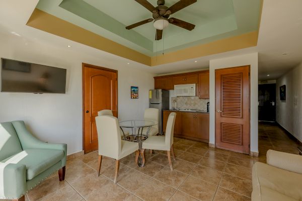 A modern living area with a TV, green armchair, glass table with four chairs, ceiling fan, and adjacent kitchenette featuring wooden cabinets.