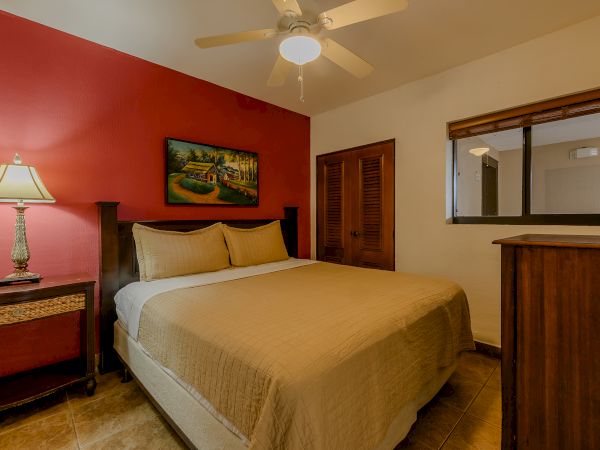 A bedroom with a double bed, two side tables, a ceiling fan, window, painting, and a dresser with a decorative item on it.