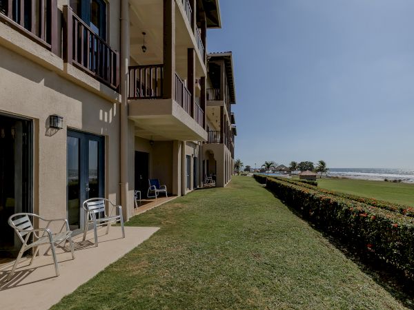 A row of beachfront apartments with balconies, patio chairs, a grassy yard, and a hedge, facing a clear sky and sea view.