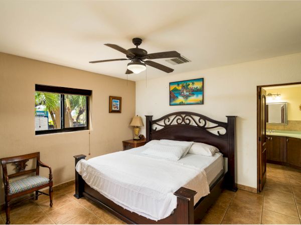 A bedroom with a double bed, wooden headboard, chair, window, ceiling fan, artwork on the wall, and an open door leading to a bathroom.