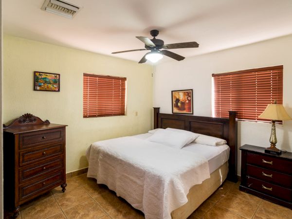 A bedroom with a double bed, wooden furniture, two paintings, and a ceiling fan. It has tiled flooring and windows with pink blinds.
