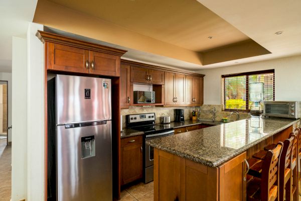 A modern kitchen with wooden cabinets, a stainless steel refrigerator, a stove, a microwave, and a granite countertop with bar stools.