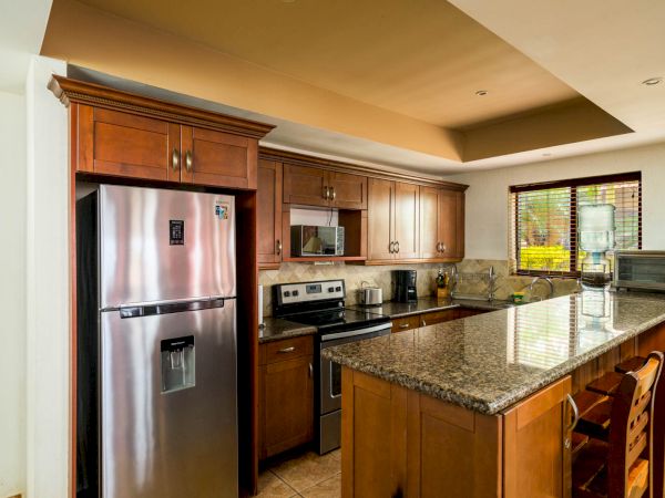 A modern kitchen with wooden cabinets, a stainless steel refrigerator, a stove, a microwave, and a granite countertop with bar stools.