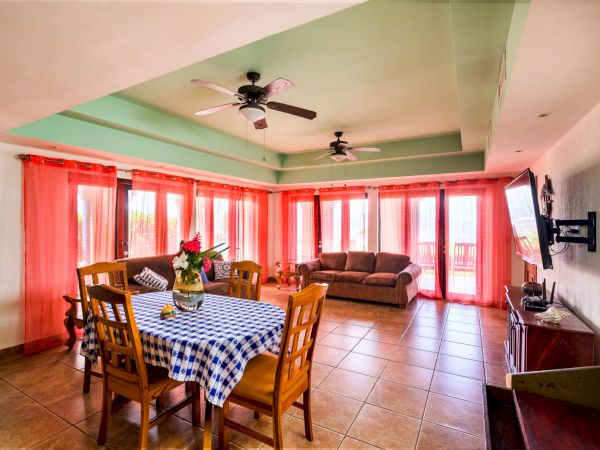 A spacious living and dining area with a checkered table, chairs, a sofa, ceiling fans, a wall-mounted TV, and large windows with orange curtains ending the sentence.
