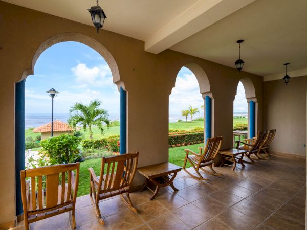 The image shows a covered patio with wooden chairs facing a beautiful outdoor landscape featuring arches, palm trees, and an ocean view in the distance.