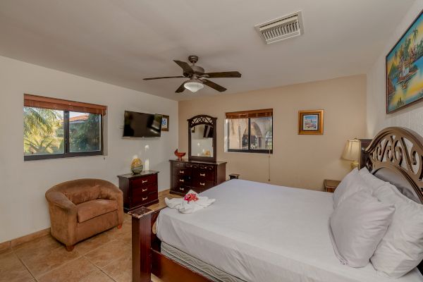 The image depicts a cozy bedroom with a large bed, wooden furniture, TV, ceiling fan, and windows. There is a chair and framed pictures on the walls.