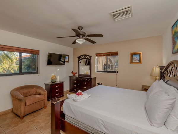 The image depicts a cozy bedroom with a large bed, wooden furniture, TV, ceiling fan, and windows. There is a chair and framed pictures on the walls.