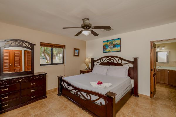 A bedroom with a wooden bed, dresser, mirror, ceiling fan, bedside lamp, and a bathroom entrance. There’s a window and pictures on the walls.