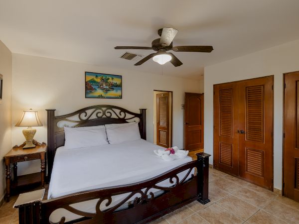 This image shows a bedroom with a large bed, a ceiling fan, a side table with a lamp, artwork on the wall, and two wooden closets.