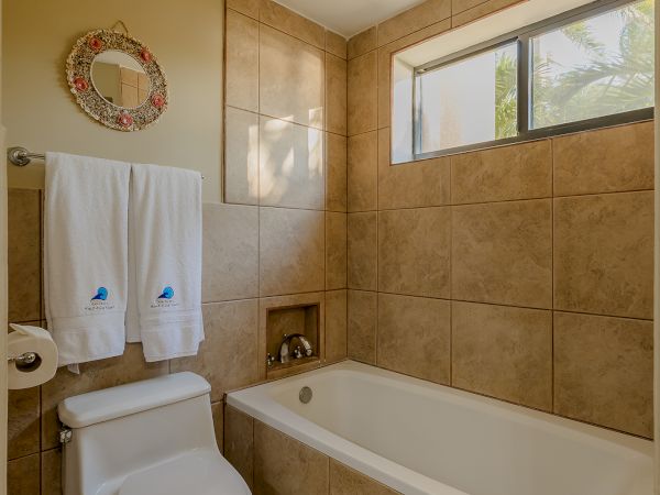 This image shows a bathroom with a bathtub, toilet, towel rack, and window. The walls and floor are tiled, and there are two white towels hanging.