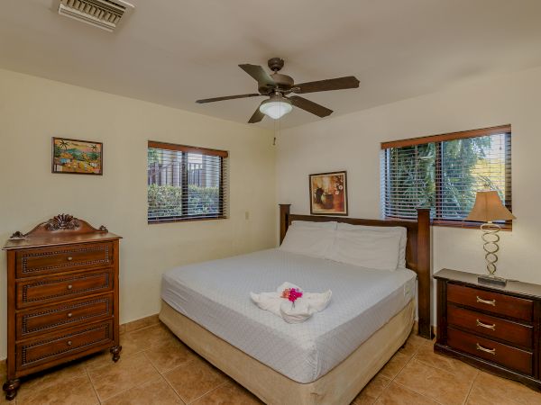 A bedroom with a double bed, two nightstands, a dresser, lamps, ceiling fan, two windows, and framed art on the walls.