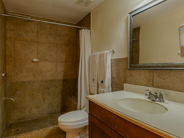 A bathroom with a shower, toilet, wooden vanity with sink, mirror, light fixture, and towels on the rack, all with a beige tile floor.