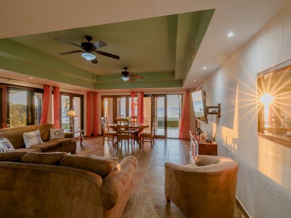 This image shows a cozy living and dining area with comfortable seating, a table, ceiling fans, and a large window with a view of the outdoors.