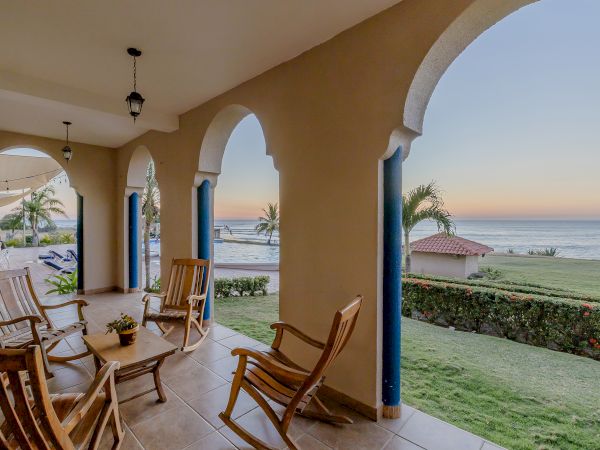 A porch with arched openings, wooden rocking chairs, and a view of a garden leading to the beach and ocean at sunset ending the sentence.