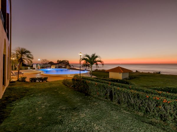 A serene coastal scene at sunset with a pool, palm trees, a small pavilion, and well-manicured lawns overlooking the calm ocean.