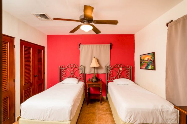 A bedroom with two single beds, a ceiling fan, a lamp on a nightstand, a painting, and wooden closet doors.