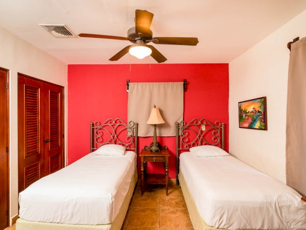 A bedroom with two single beds, a ceiling fan, a lamp on a nightstand, a painting, and wooden closet doors.