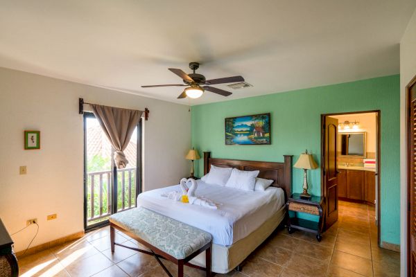 A cozy bedroom with a large bed, ceiling fan, wooden furniture, decorative picture, bench at the foot of the bed, and a window partially covered by a curtain.