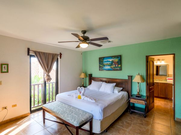 A cozy bedroom with a large bed, ceiling fan, wooden furniture, decorative picture, bench at the foot of the bed, and a window partially covered by a curtain.