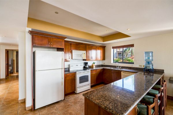 A modern kitchen with wooden cabinets, granite countertops, a white refrigerator, stove, microwave, and a water dispenser on a long island.