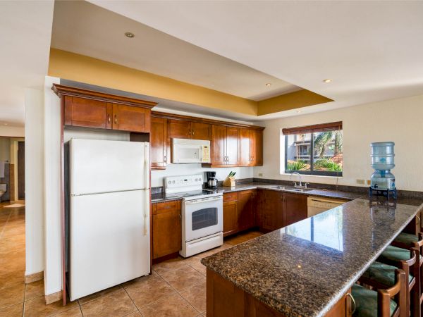 A modern kitchen with wooden cabinets, granite countertops, a white refrigerator, stove, microwave, and a water dispenser on a long island.