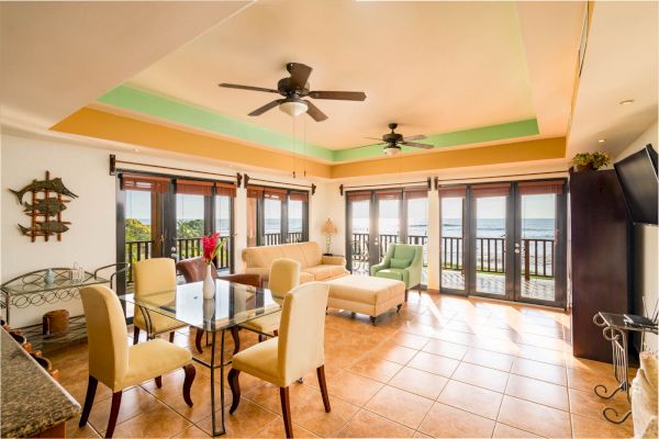 The image shows a bright, open living and dining area with large windows, ceiling fans, and modern furnishings, overlooking a balcony with a scenic view.