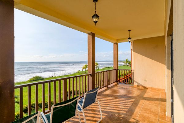 A beachfront patio with a scenic ocean view, wooden railings, two chairs, tile flooring, and hanging lanterns on the ceiling, bathed in sunlight.