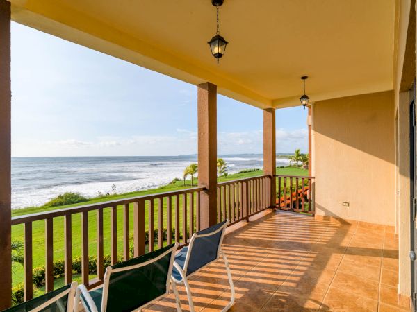 A beachfront patio with a scenic ocean view, wooden railings, two chairs, tile flooring, and hanging lanterns on the ceiling, bathed in sunlight.