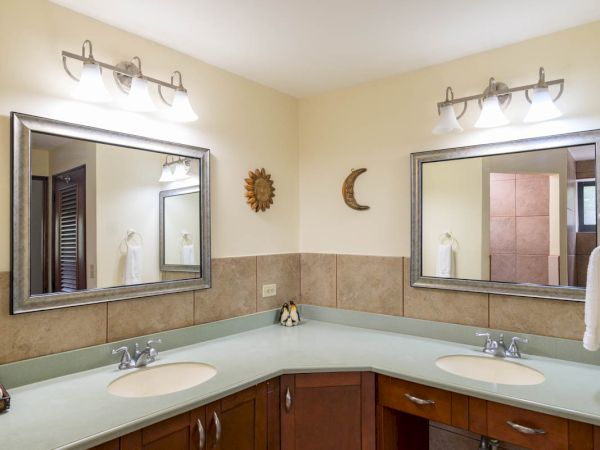 This image shows a bathroom with a double sink vanity, two mirrors, overhead lighting, wooden cabinets, and wall decor under warm lighting.