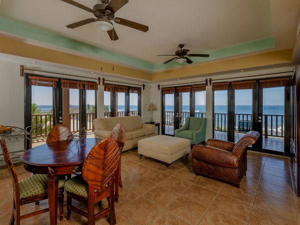 A spacious living room with multiple windows offering an ocean view, comfortable seating, ceiling fans, a dining table, and tiled flooring.