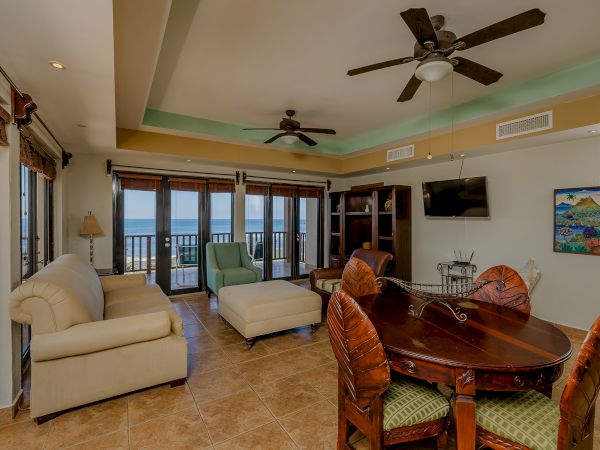 A spacious living room with beige furnishings, a dining table with chairs, large windows, ceiling fans, a wall-mounted TV, and an ocean view ends the sentence.