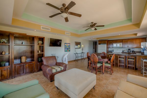 A spacious living room with two ceiling fans, a kitchen, an island with stools, a dining area, and cozy seating facing a TV.