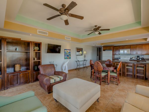 A spacious living room with two ceiling fans, a kitchen, an island with stools, a dining area, and cozy seating facing a TV.