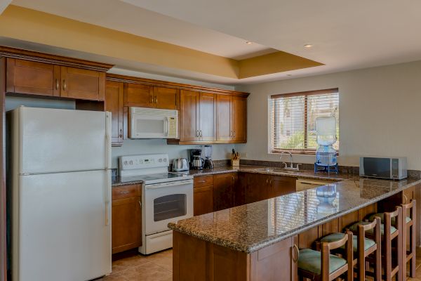 A modern kitchen with wooden cabinets, granite countertops, white appliances, a bar with stools, and a window providing natural light.