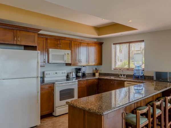 A modern kitchen with wooden cabinets, granite countertops, white appliances, a bar with stools, and a window providing natural light.