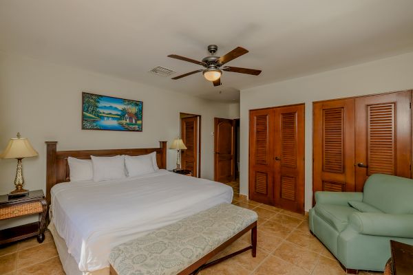 A cozy bedroom features a king-size bed, a green armchair, wooden furniture, a ceiling fan, and a landscape painting on the wall.